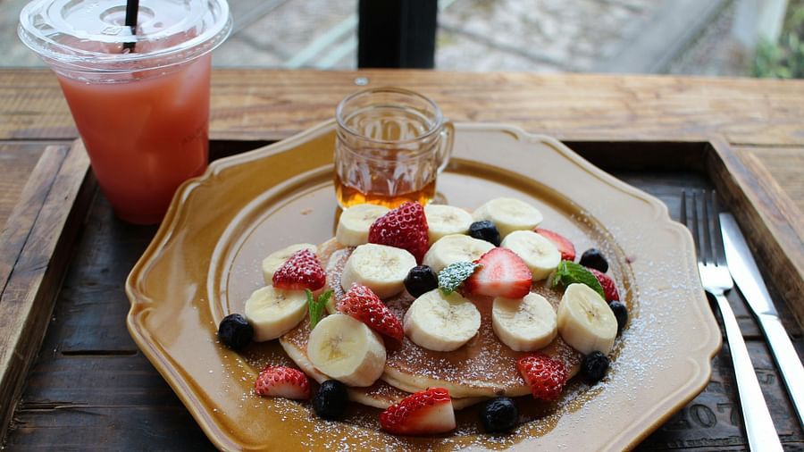 A plate of pancakes topped with banana slices, strawberries, blueberries, and powdered sugar, served with syrup and a glass of pink juice. 