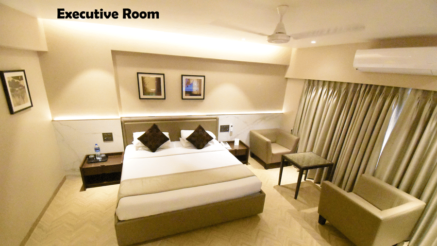 An Executive Room at Kings International Juhu featuring a bed, 2 sofas and a centre table, an A/C unit, and paintings on the walls.