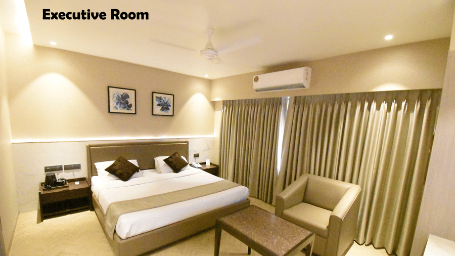 An Executive Room at Kings International Juhu with a neatly-made bed, a sitting area, an A/C unit, and paintings on the wall.
