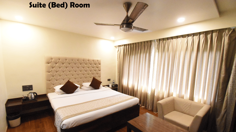 The bedroom of a Suite Room featuring a bed with a tufted headboard, a nightstand, a ceiling fan and full-length curtains | Kings International Juhu