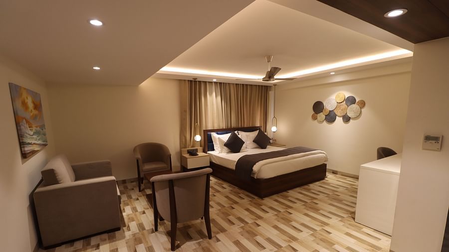 Side angle view of a Cozy bed with bed side tables, sofa set with coffee table and painting hanging over it and ceiling fan in  studio bedroom - Mastiff Hotel, Dhanbad