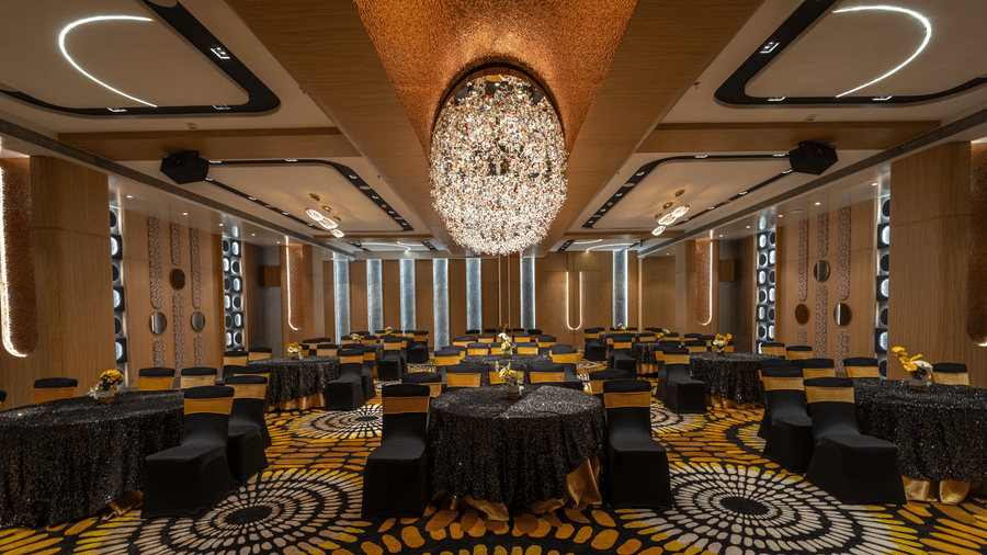 A banquet hall featuring round tables with black table cloths, yellow and black patterned carpet, and a large, linear chandelier | Pride Elite Chhatrapati Sambhajinagar