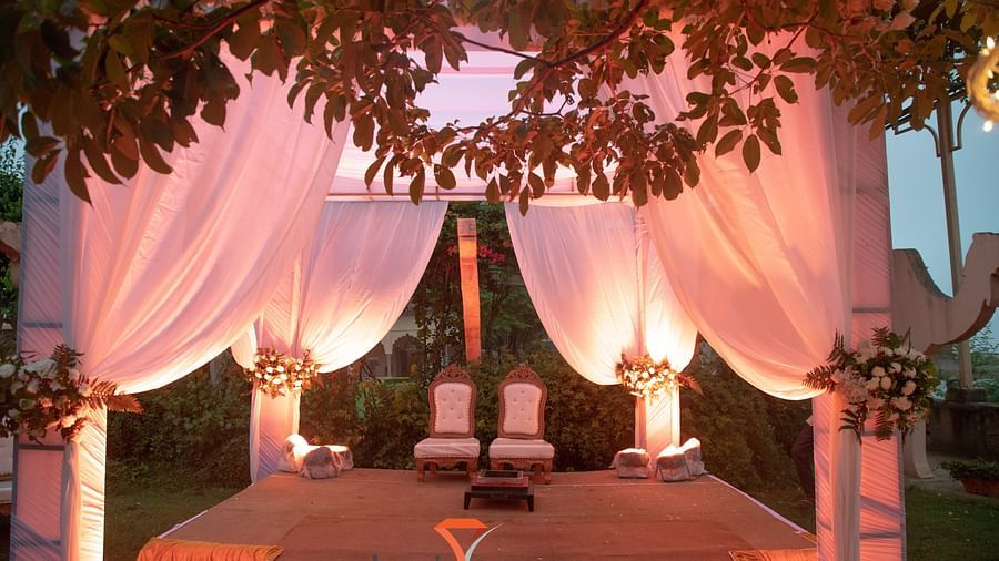 A decorated stage with chairs and draped white fabric is prepared for an event at Tijara Fort-Palace - 19th Century, Alwar, under the branches of a tree.