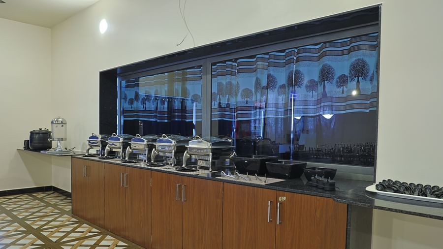 A buffet counter at the VITS Select Diamond Resort, Sasan Gir restaurant. The counter has several steel serving dishes and bowls, with a large window and patterned curtains behind them.