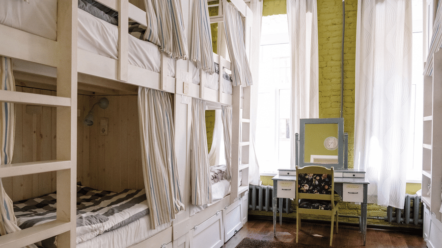 A room containing rows of wooden bunk beds with curtains, next to a small desk and chair by a window.