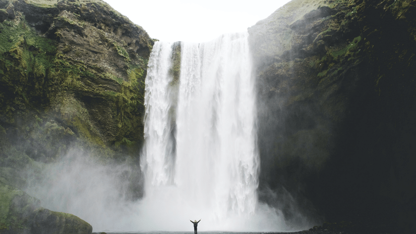 A waterfall during the day and a person standing while facing it with their open arms