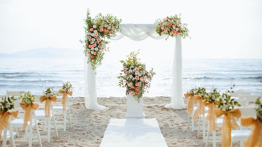 Beachside wedding set up featuring a stage decorated with flowers and fabric pieces. At the centre there is a aisle and on either side there are chairs decorated for guests.