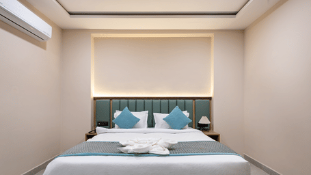 A king bed with white linens and blue accent pillows, 2 side tables, and an air-conditioner in a guest room at Monday Hotels & Suites Madhapur Hyderabad.
