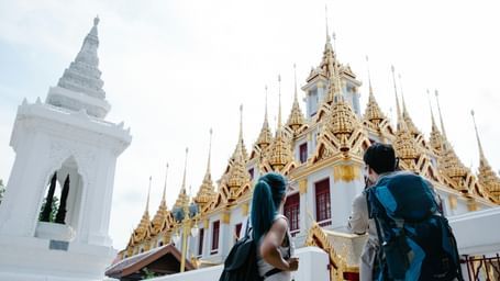 Two tourists with backpacks view a golden-roofed temple in Thailand, with an ornate white structure in the foreground.