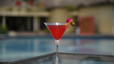 A close-up of a red martini cocktail glass placed on the edge of the swimming pool at Vedic Village Spa Resort with a blurred pool background in a warm outdoor setting.