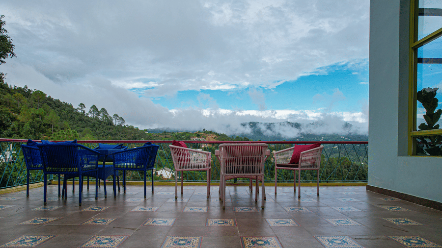An outdoor terrace with chairs and tables placed on tiled flooring facing hills and sky at Adrushya Estates, Mukteshwar.