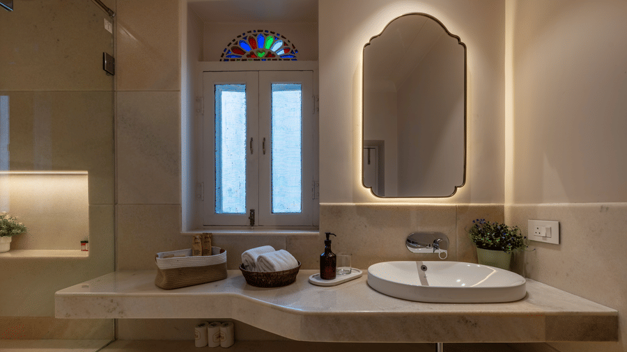 A bathroom vanity at Barwara Kothi, Jaipur featuring a marble counter, basin, illuminated mirror, basket, and stained glass window.