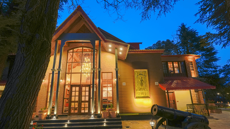 The well-lit facade of BluSalzz Terrah Hills Resort, Dalhousie, with steps in the entrance, a large wooden door, and trees in the backdrop at night.