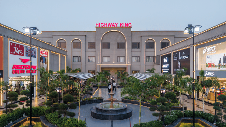A central circular water fountain, manicured green lawns with palm trees, and retail storefronts under lights at Hotel Highway King, Harmada.