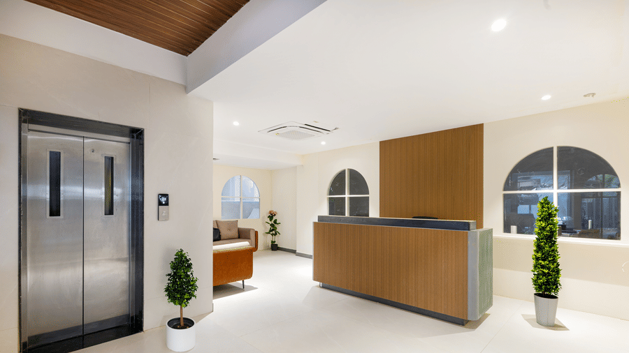 An elevator with stainless steel doors and a wooden reception desk stand in a spacious white lobby area.