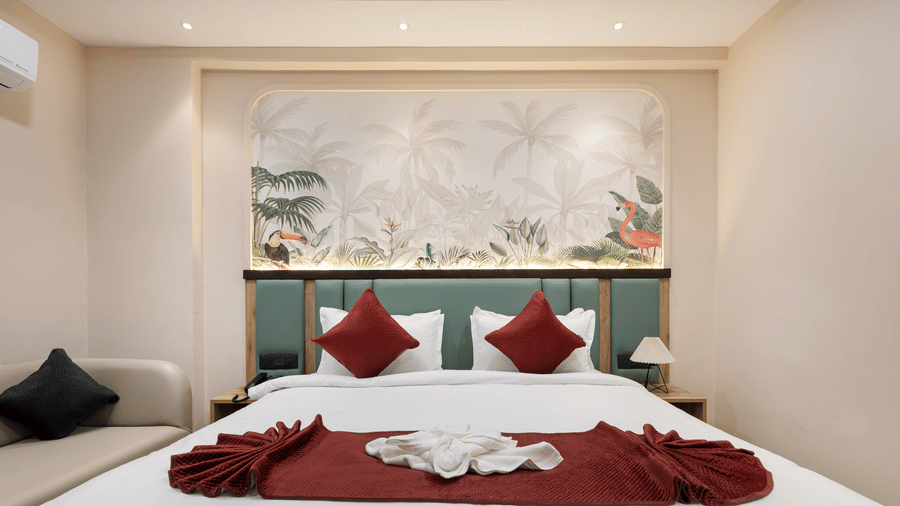 A bedroom showing a king size bed with red decorative pillows and a dark red bed runner, a sofa, and an air-conditioner, at Monday Hotels & Suites Madhapur Hyderabad.