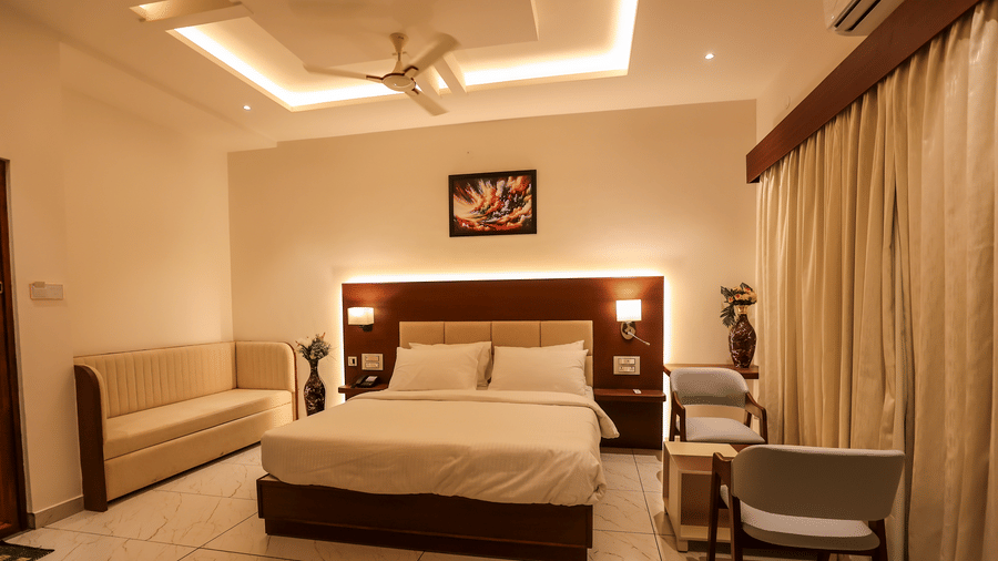 A well-lit hotel bedroom featuring a double bed, a beige sofa against the wall, an illuminated headboard, and a desk chair | Lords Sahyadri Resort, Shivamogga