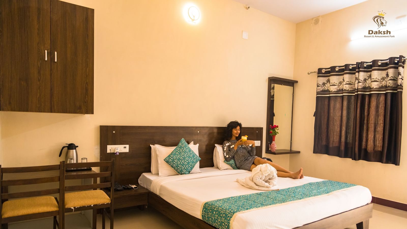 A guest room with a double bed, chairs, and dressing area, with a woman seated at Daksh Resort & Amusement Park.