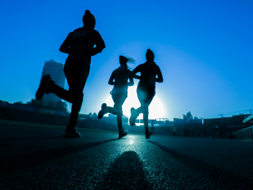 Three runners silhouetted against a bright sky and low sun on an open road
