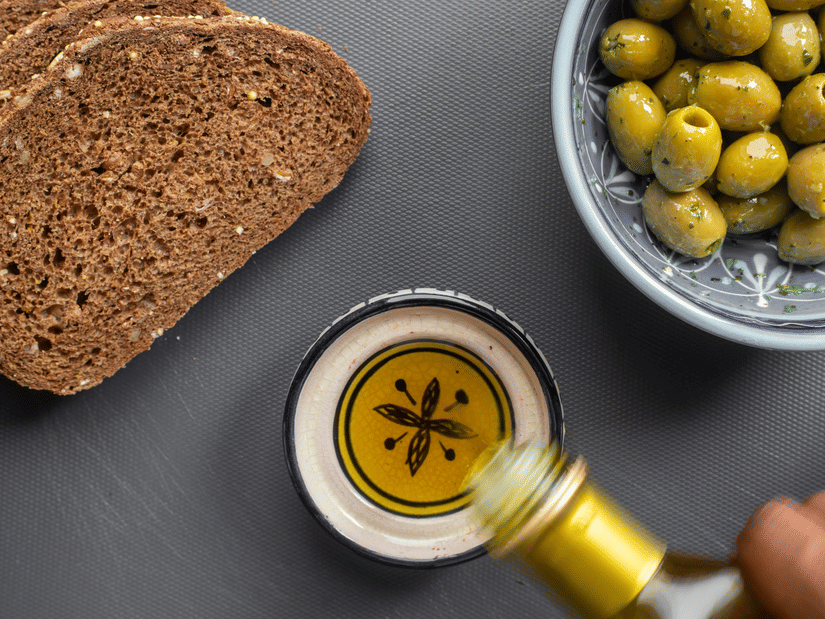 Two slices of dark rye bread, a bowl of green olives and a glass carafe of olive oil on a dark surface