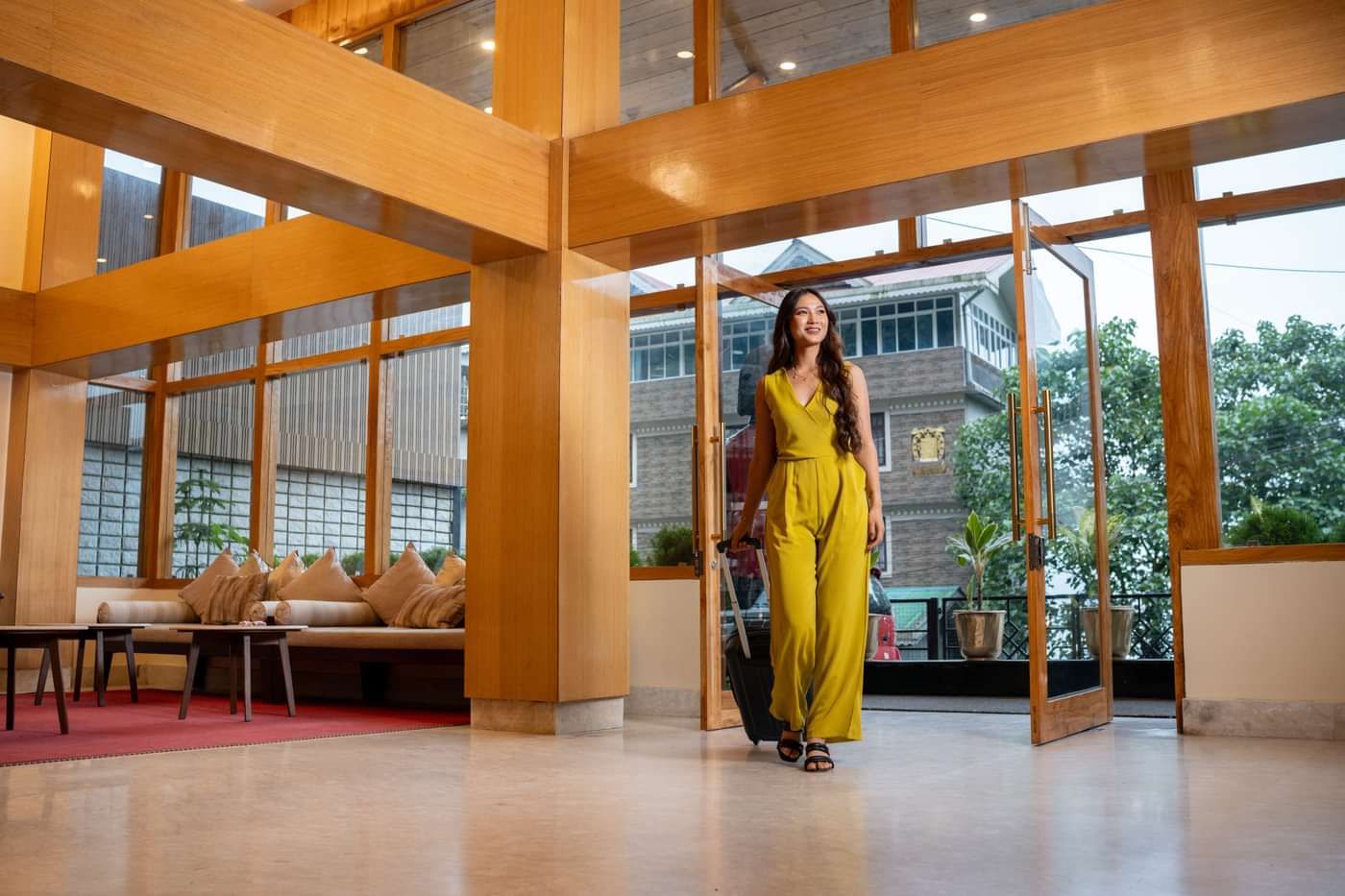 A woman in a yellow jumpsuit walks into a hotel lobby with a suitcase, passing through glass doors. The lobby has wooden frames, seating, and outside greenery.