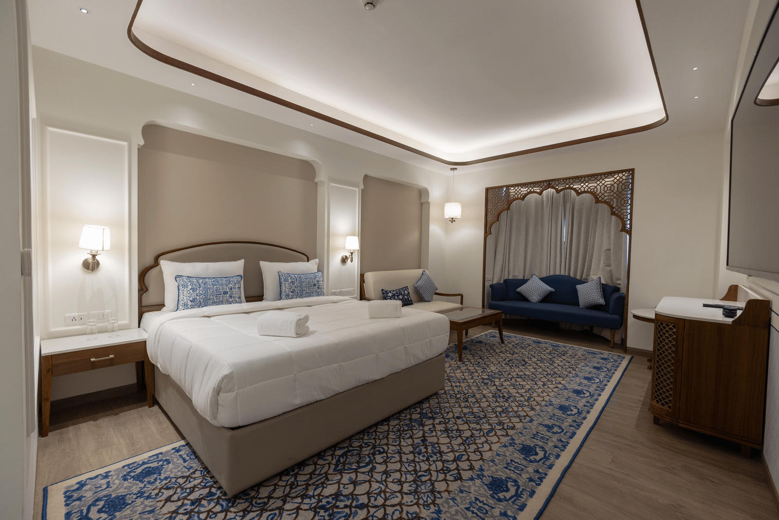 A warmly lit bedroom of Mandakini at Bhagirathi by Atishay featuring a plush bed with lamps on the wall of either side of it and seating area by the window with closed curtains