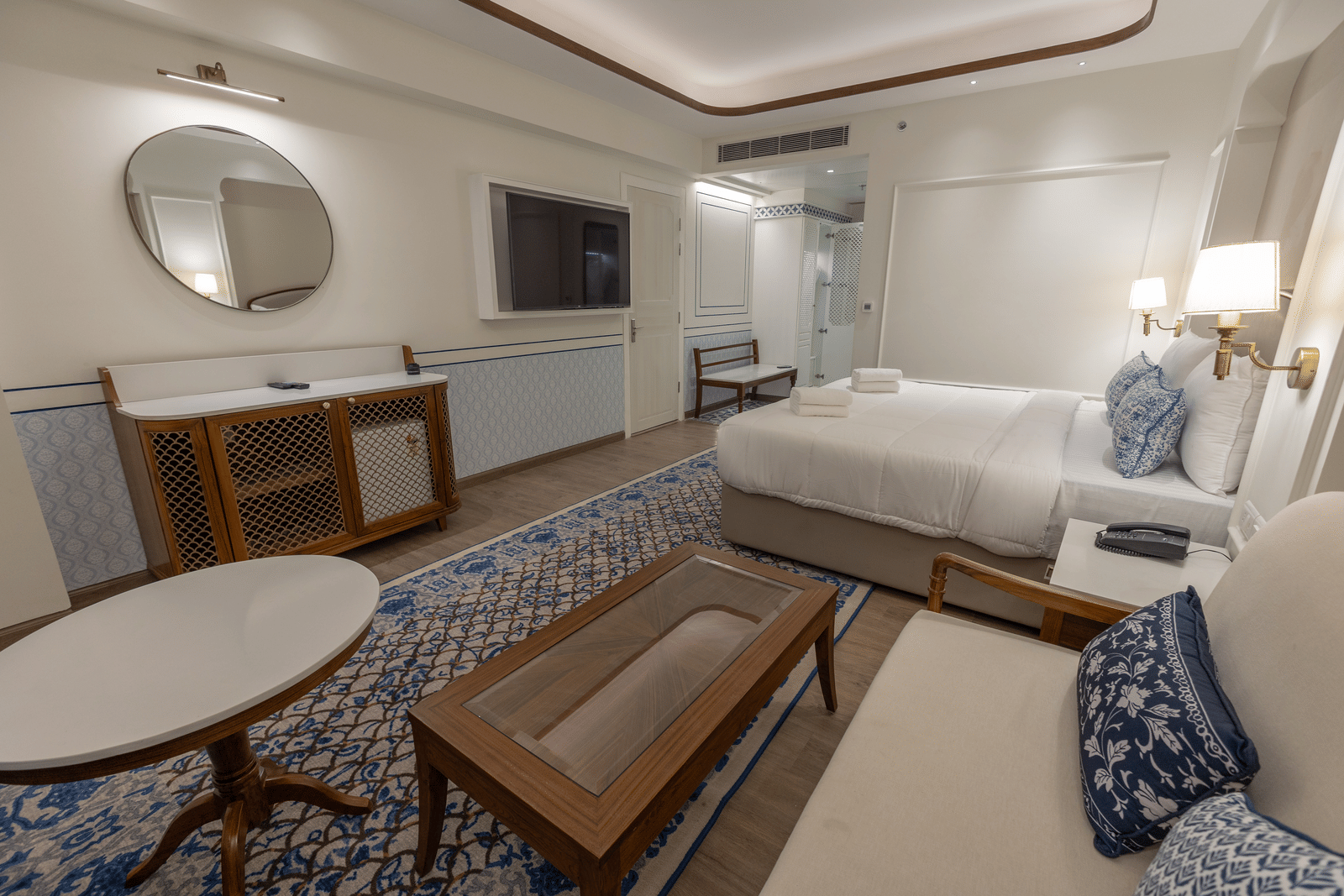 A warmly lit Triveni bedroom featuring a plush bed with two rolled towels placed on it facing the TV on the wall and seating space with coffee table next to the nightstand at Bhagirathi by Atishay