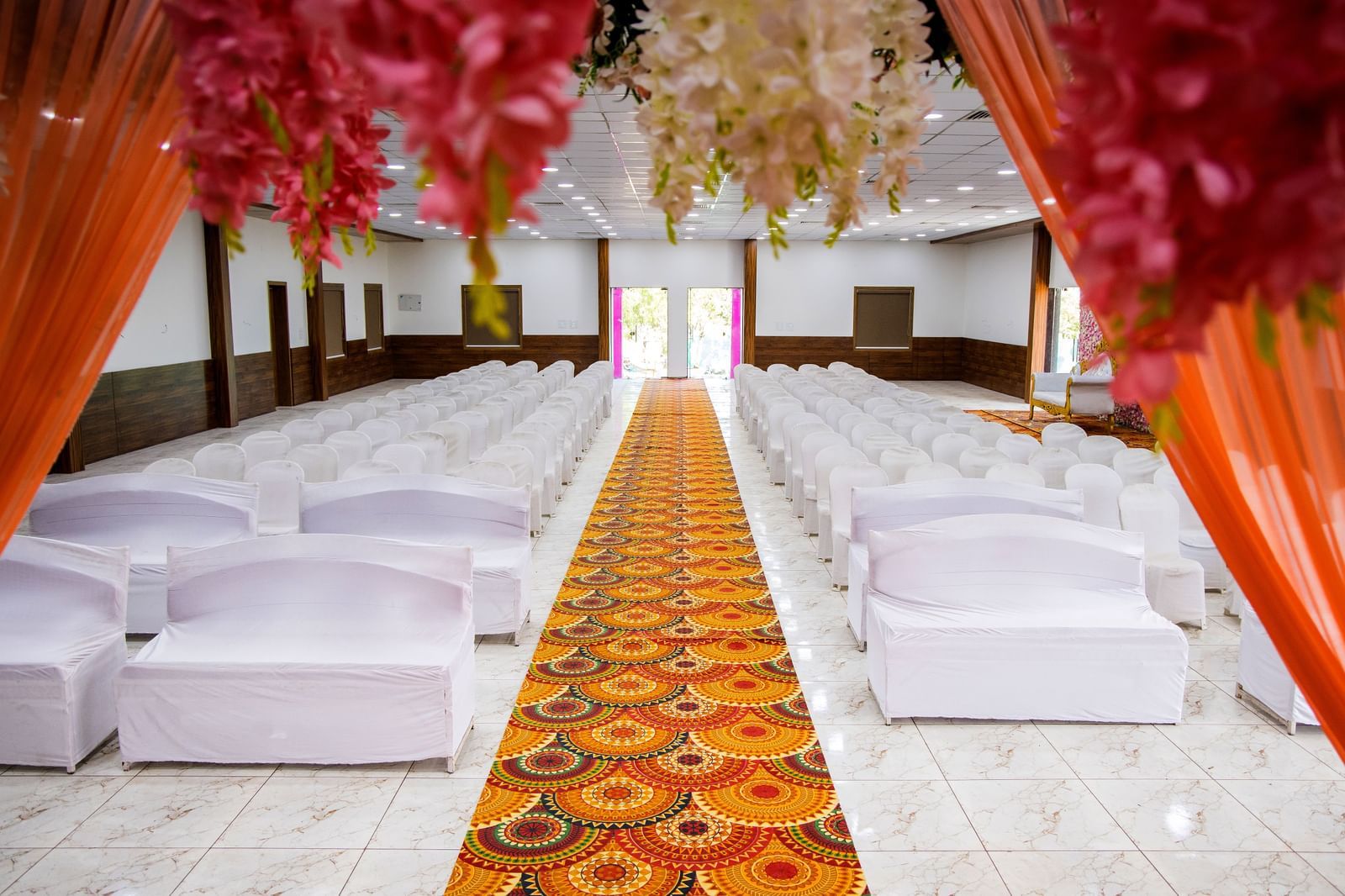 A close up of a hall at Daksh Resort & Amusement Park featuring arranged chairs and a patterned isle with decorated curtains on the foreground.