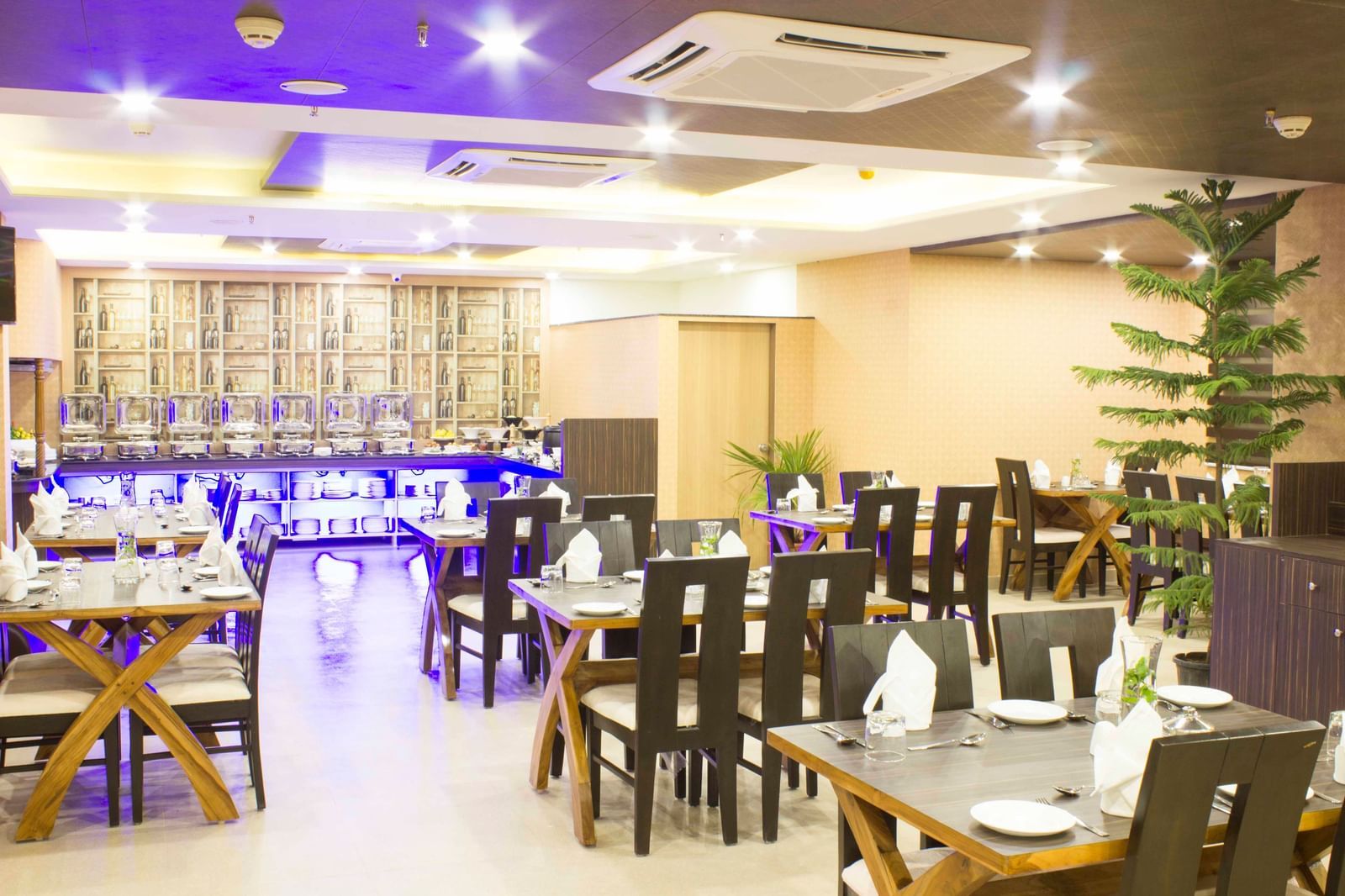 A hotel restaurant with set tables, dark chairs, and blue ambient lighting | Daksh The Madhuvan Suites