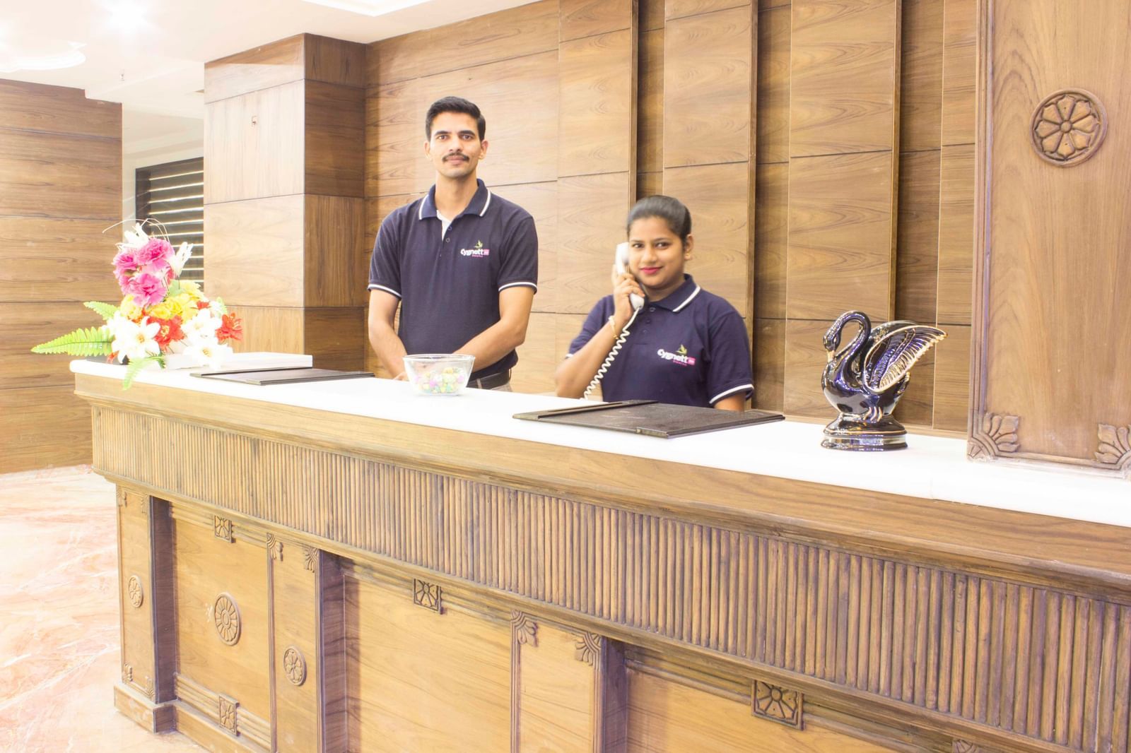 A hotel reception desk with two staff members smiling | Daksh The Madhuvan Suites