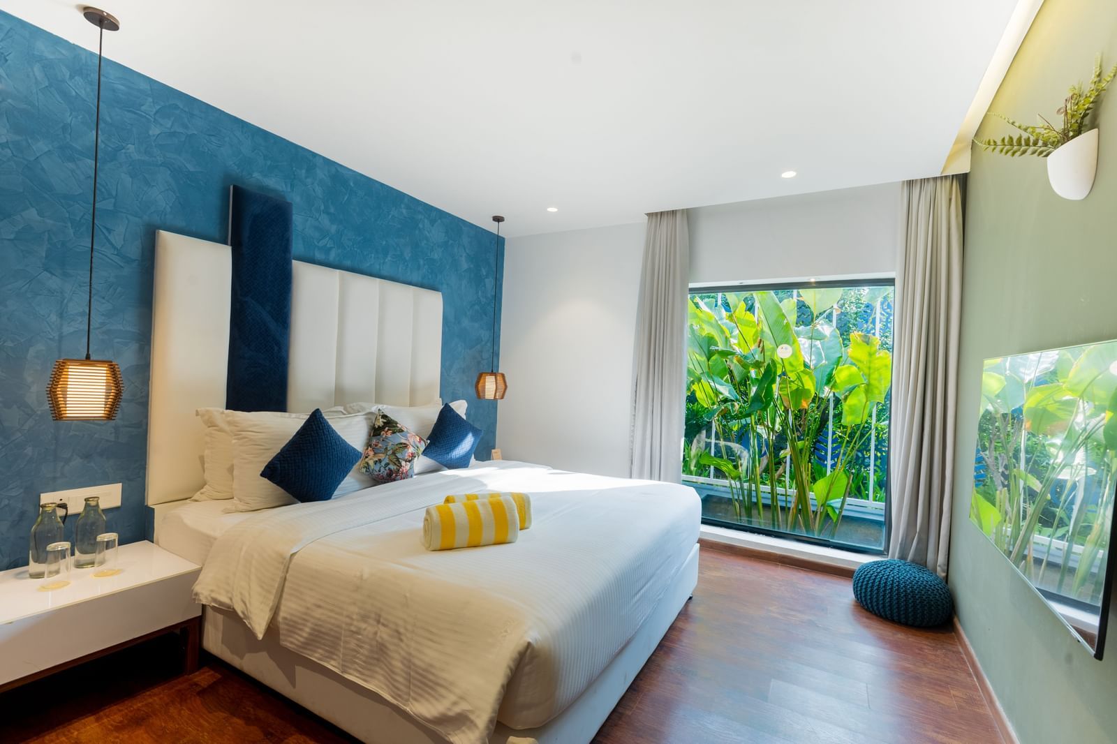 Bedroom of Two Bedroom Villa at Darza Resorts features a large bed, blue accent wall, and a full-length window overlooking lush tropical foliage. 