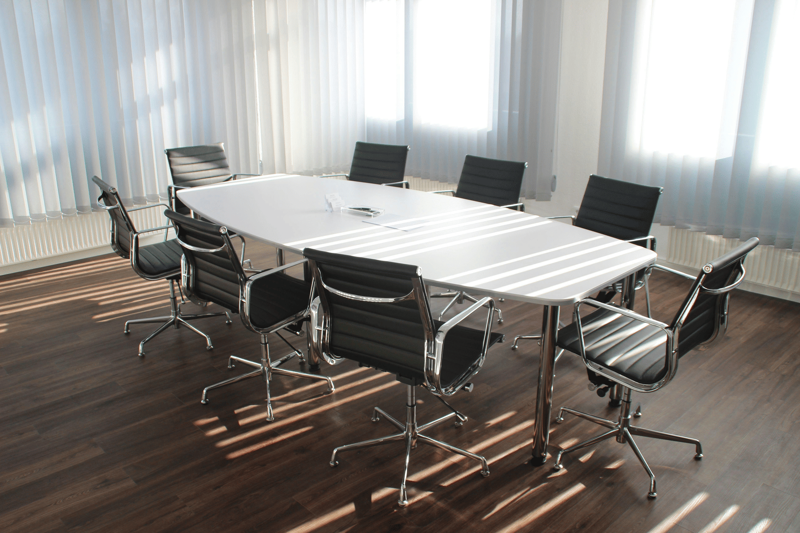 A bright and spacious meeting room with a white oval conference table, black swivel chairs, and large windows allowing natural light.