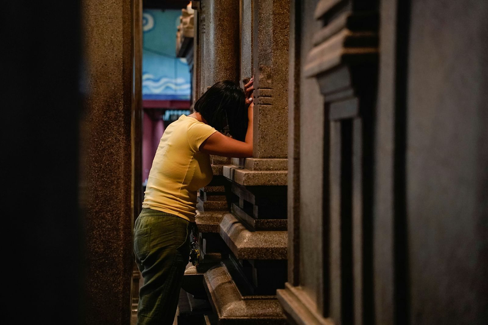 Image of a woman wearing yellow shirt leaning her head on a stoned wall in a temple