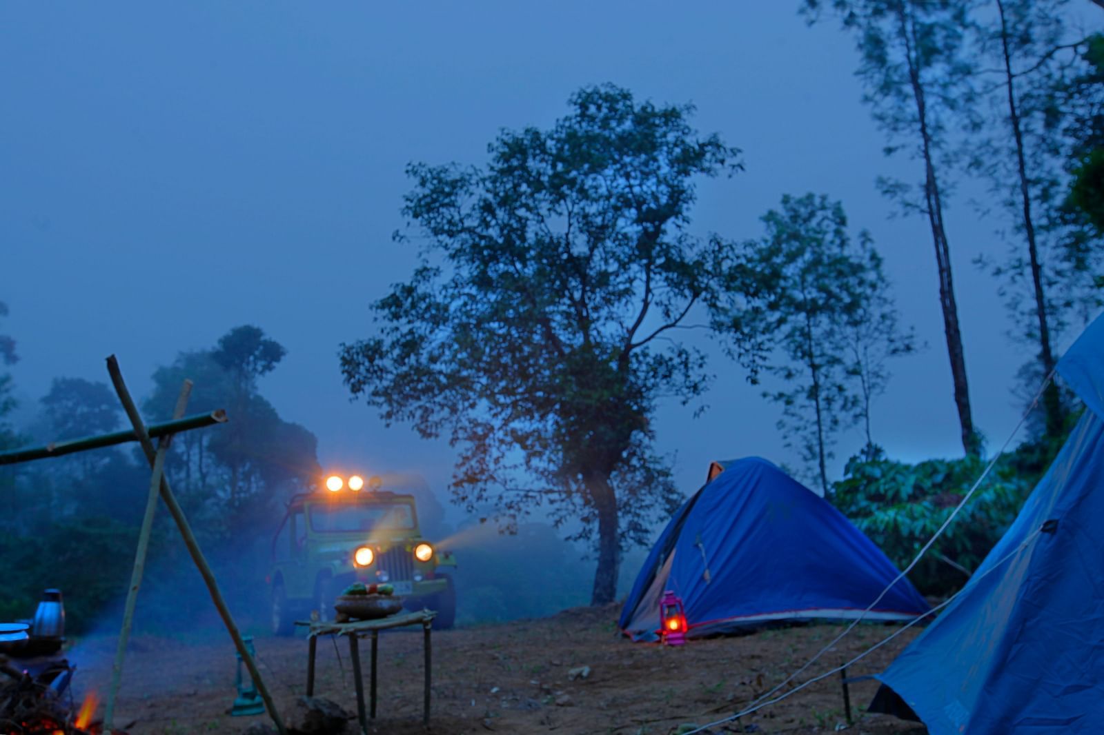 An evening campsite scene with a blue tent, a fire pit, and a vehicle with headlights on in a misty forest setting - Evergreen County