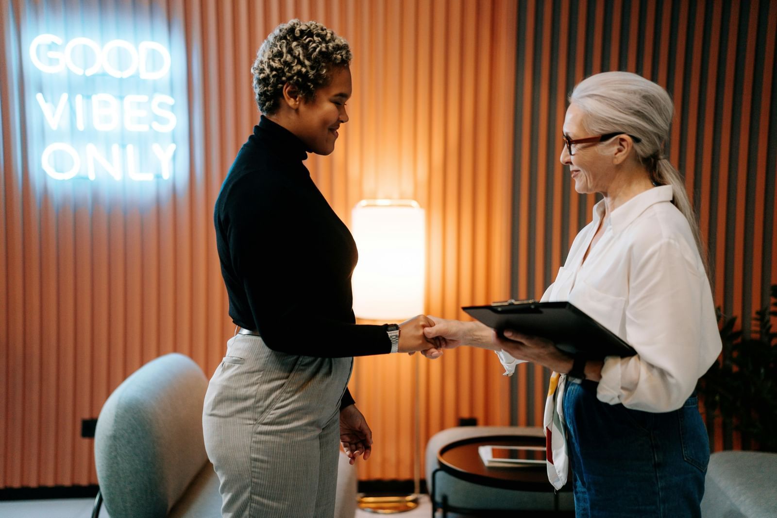 Two women in a professional setting shake hands, with one holding a folder.