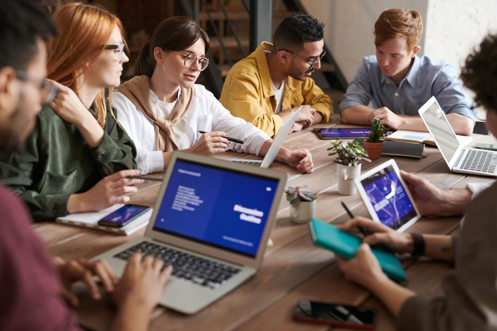 A group of people are gathered around a table, using laptops and tablets in a collaborative work environment.