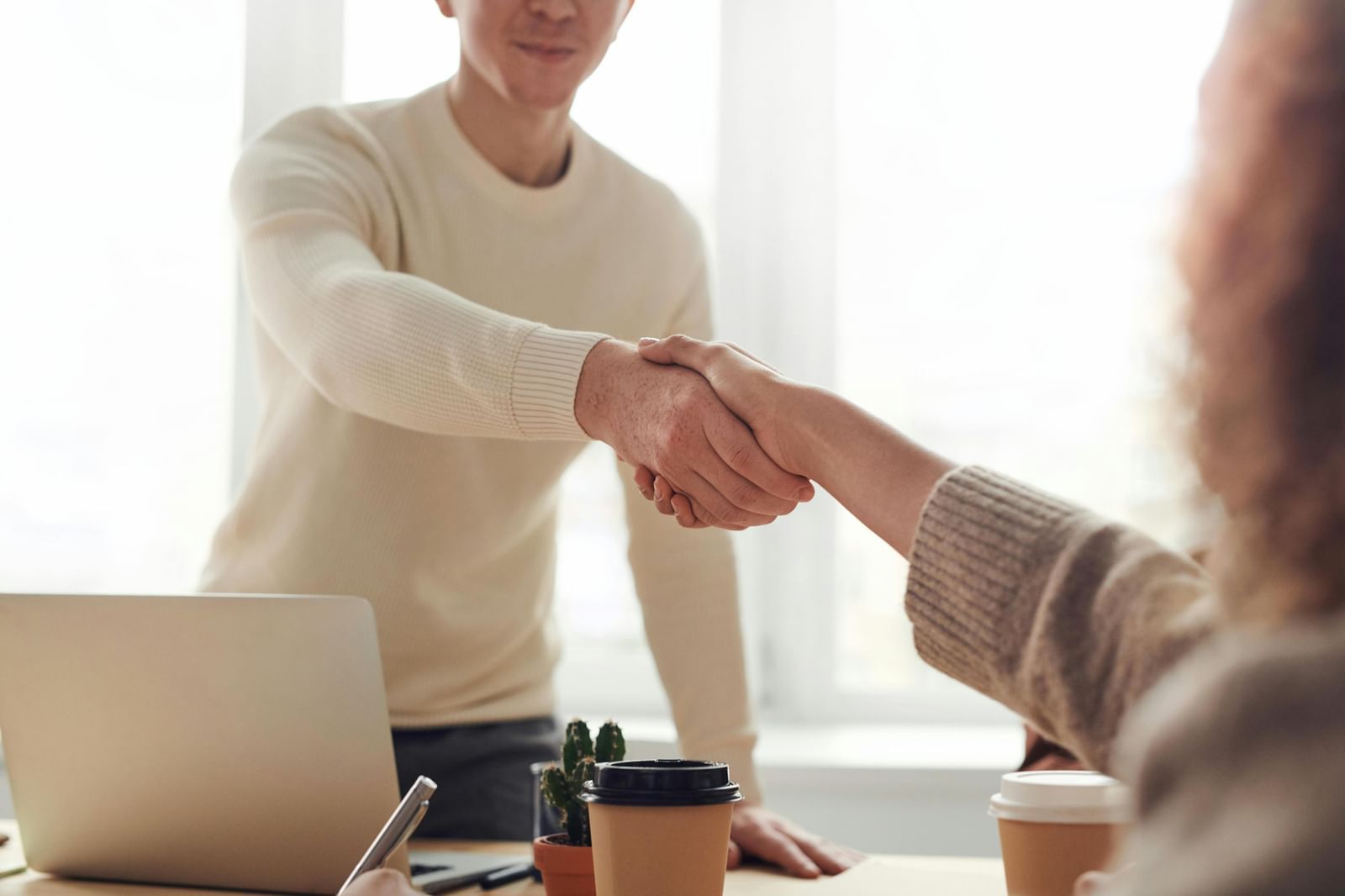 A handshake between two people in a professional setting, with a laptop and coffee cups on a desk in the foreground.