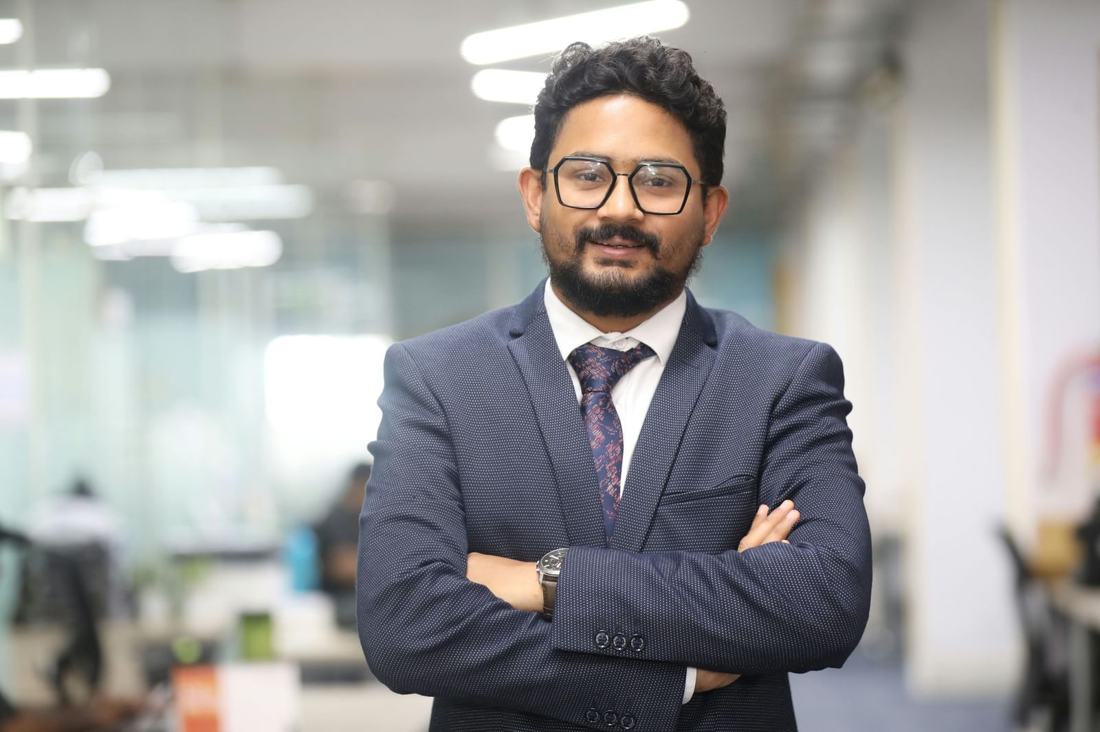 A confident business professional with glasses and a beard stands with arms crossed in an office setting.