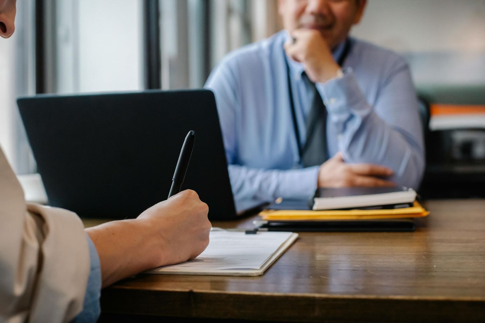A person taking notes on a laptop during a meeting or interview, with another person smiling in the background.