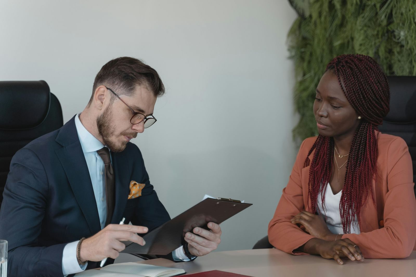 A man in a suit reviews documents while a woman in a coral blazer looks on.