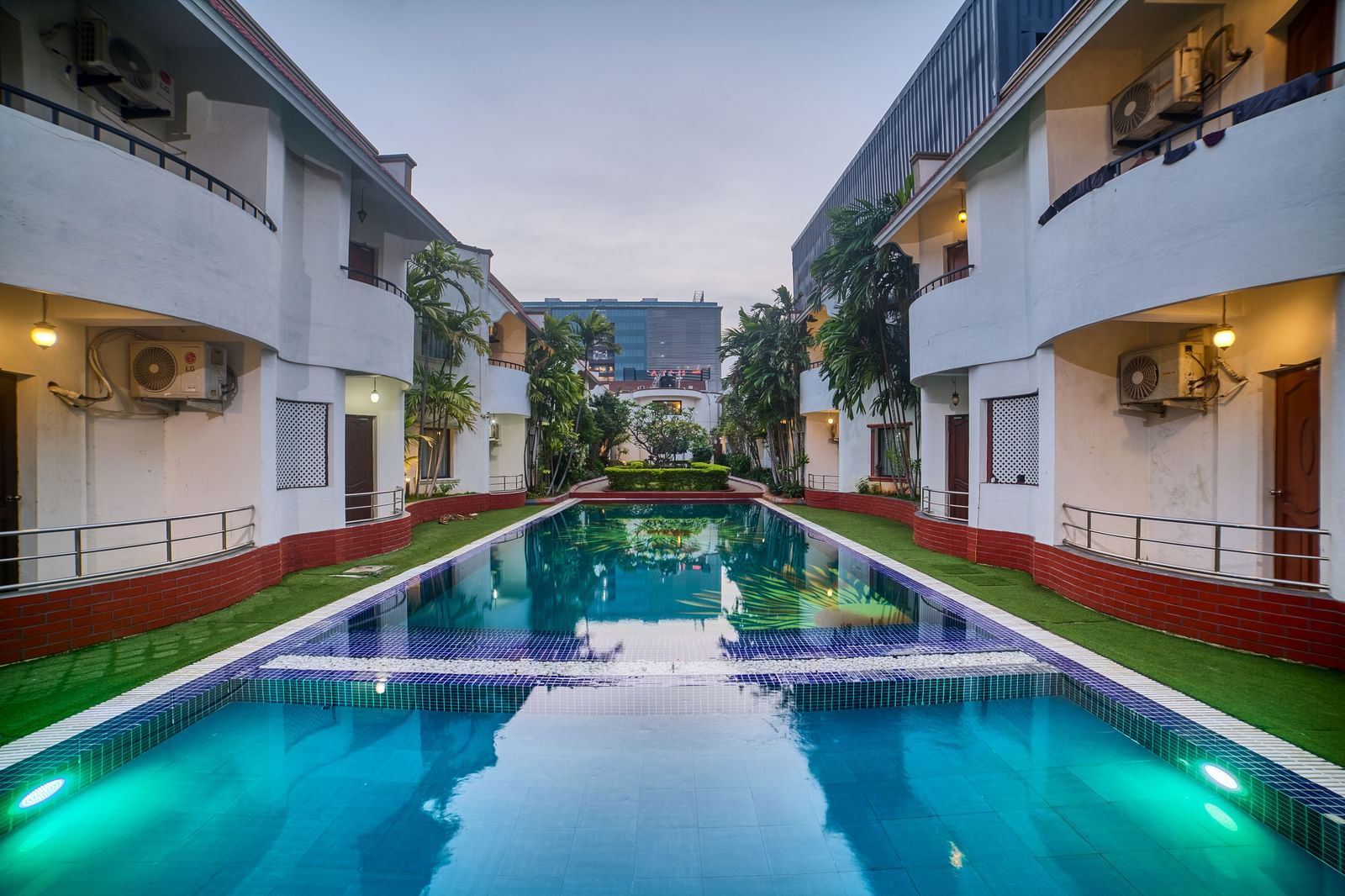A symmetrical view of Ilara Hotels & Spa, OMR, Chennai illuminated at dusk, lining a long, reflective swimming pool or water feature.