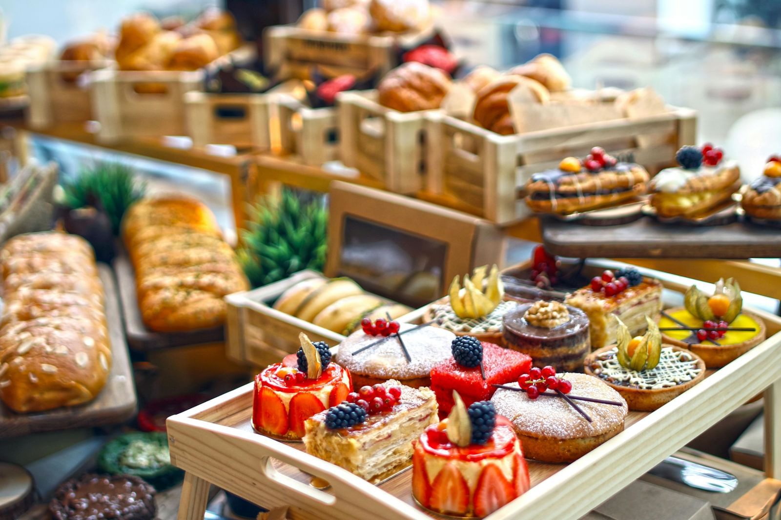 A vibrant bakery display featuring assorted pastries, cakes, tarts, loaves of bread, and eclairs arranged in wooden trays.