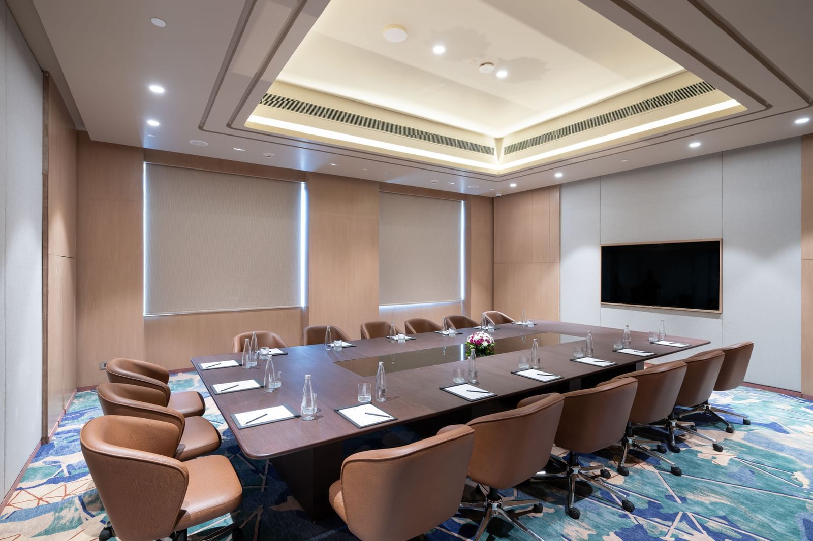 Diagonal view of a conference room featuring a projector, large table, and cushioned chairs at Merlis Hotel, Coimbatore.