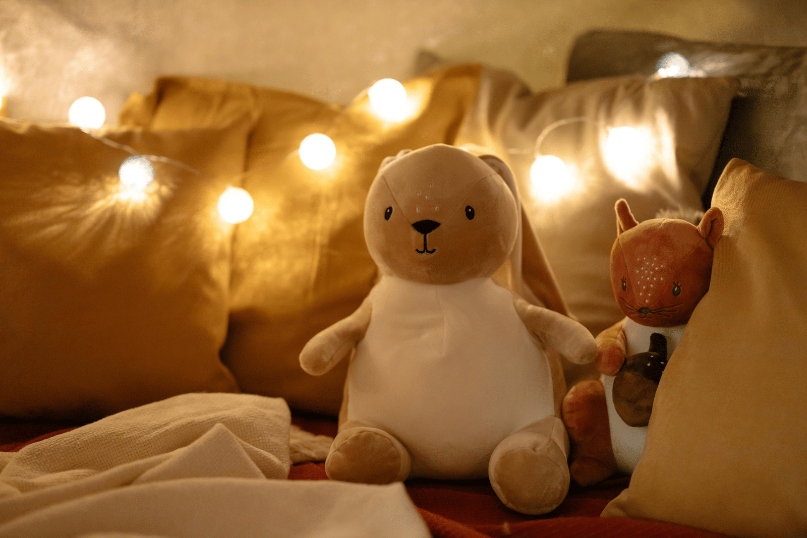 A teddy bear sits on a bed with warm fairy lights strung above.