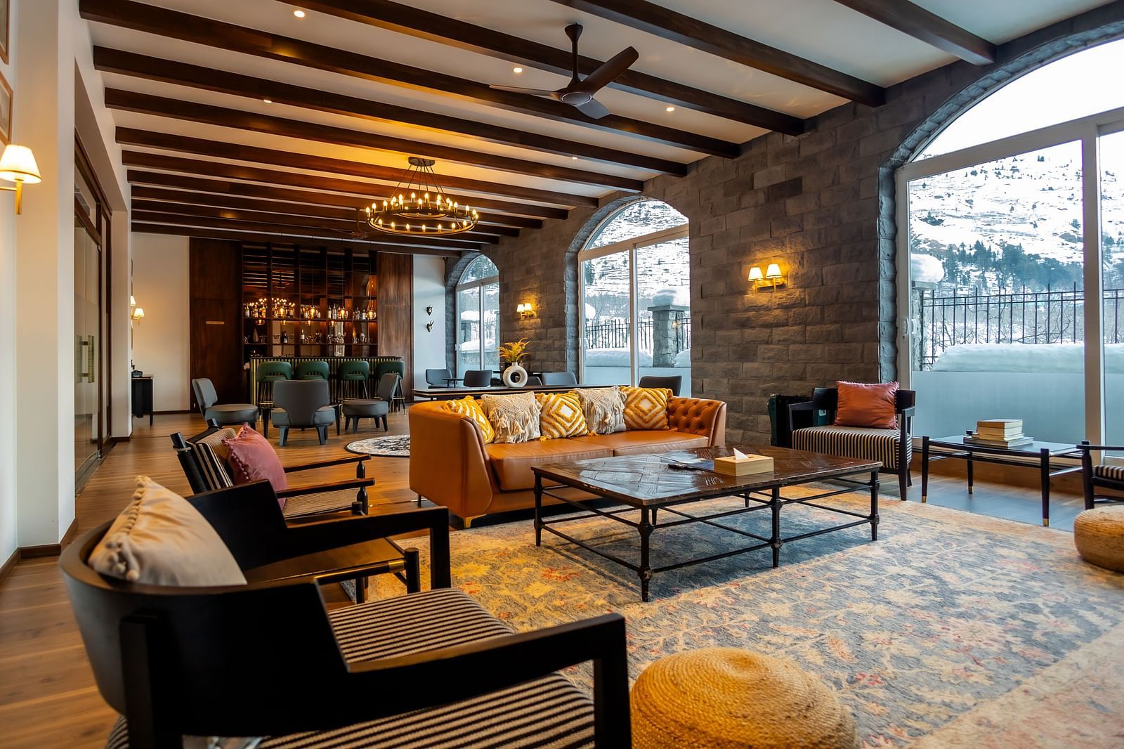 A bar boasting exposed beams in the ceiling with warm lighting and a chandelier, stone walls, plush seating, and large windows showcasing snowy mountain vistas at Nivasana Manali.