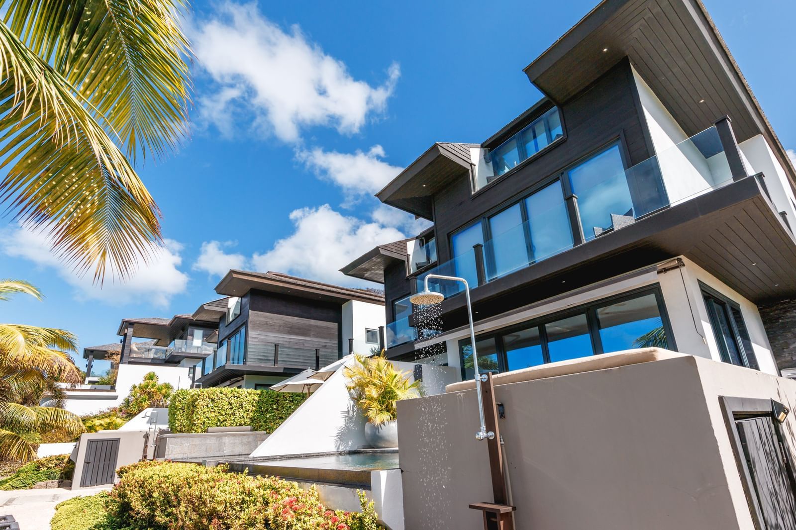 Modern, multi-story luxury villas with large windows and balconies, surrounded by lush green foliage under a sunny sky.