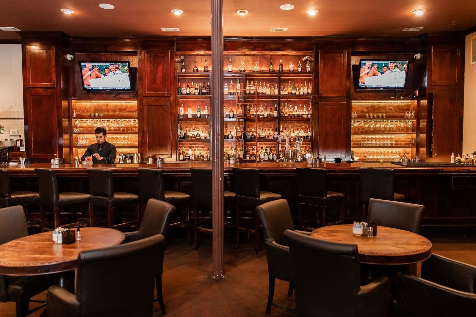 A bar area with round tables, a liquor display on the back wall, and mounted televisions above.- The Grand Hotel & Ballroom, McKinney
