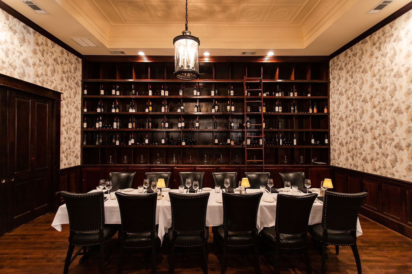 A  dining room with a long table, chairs, and a floor-to-ceiling rack of bottles against the back wall.- The Grand Hotel & Ballroom, McKinney