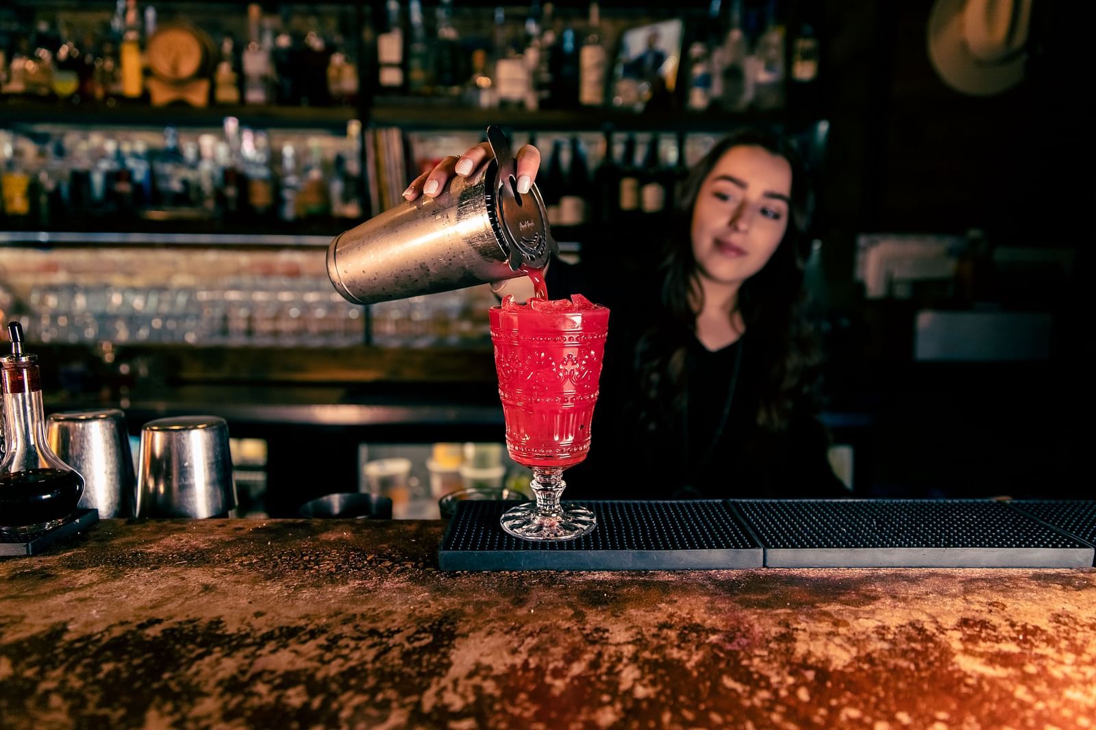 A bartender pours a drink into a glass at a bar, with shelves of liquor bottles in the background.- The Grand Hotel & Ballroom, McKinney