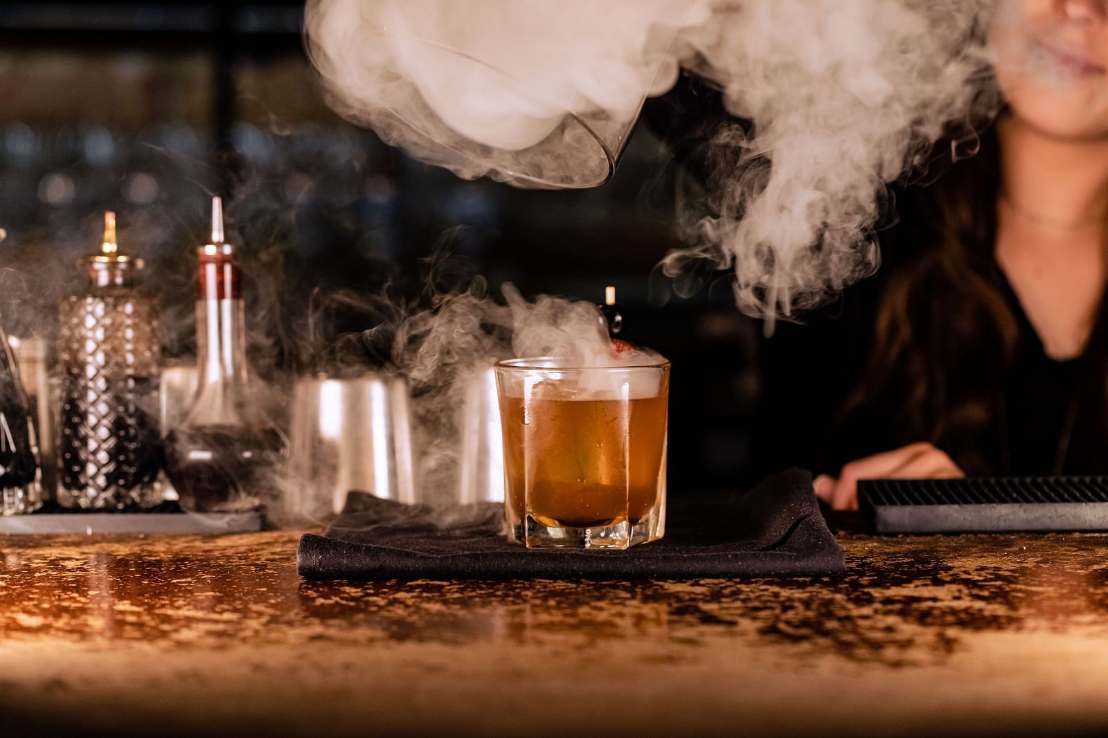 A bartender prepares a drink, with smoke rising from the drink, surrounded by bar tools and ingredients.- The Grand Hotel & Ballroom, McKinney